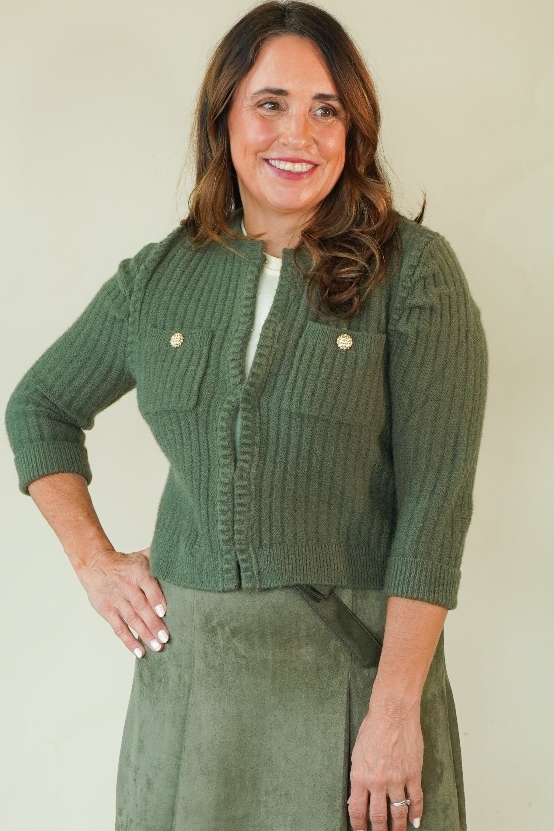 A woman modeling a matching olive green two-piece set consisting of a textured button-front cardigan and a corduroy mini skirt, paired with beige wedge sandals, against a plain cream background.