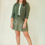 A woman modeling a matching olive green knit cardigan and skirt set, styled with a white top and beige wedge sandals, against a plain cream background.