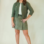 A woman modeling a matching olive green knit cardigan and skirt set with a white top and beige wedge sandals, standing against a plain cream studio backdrop.
