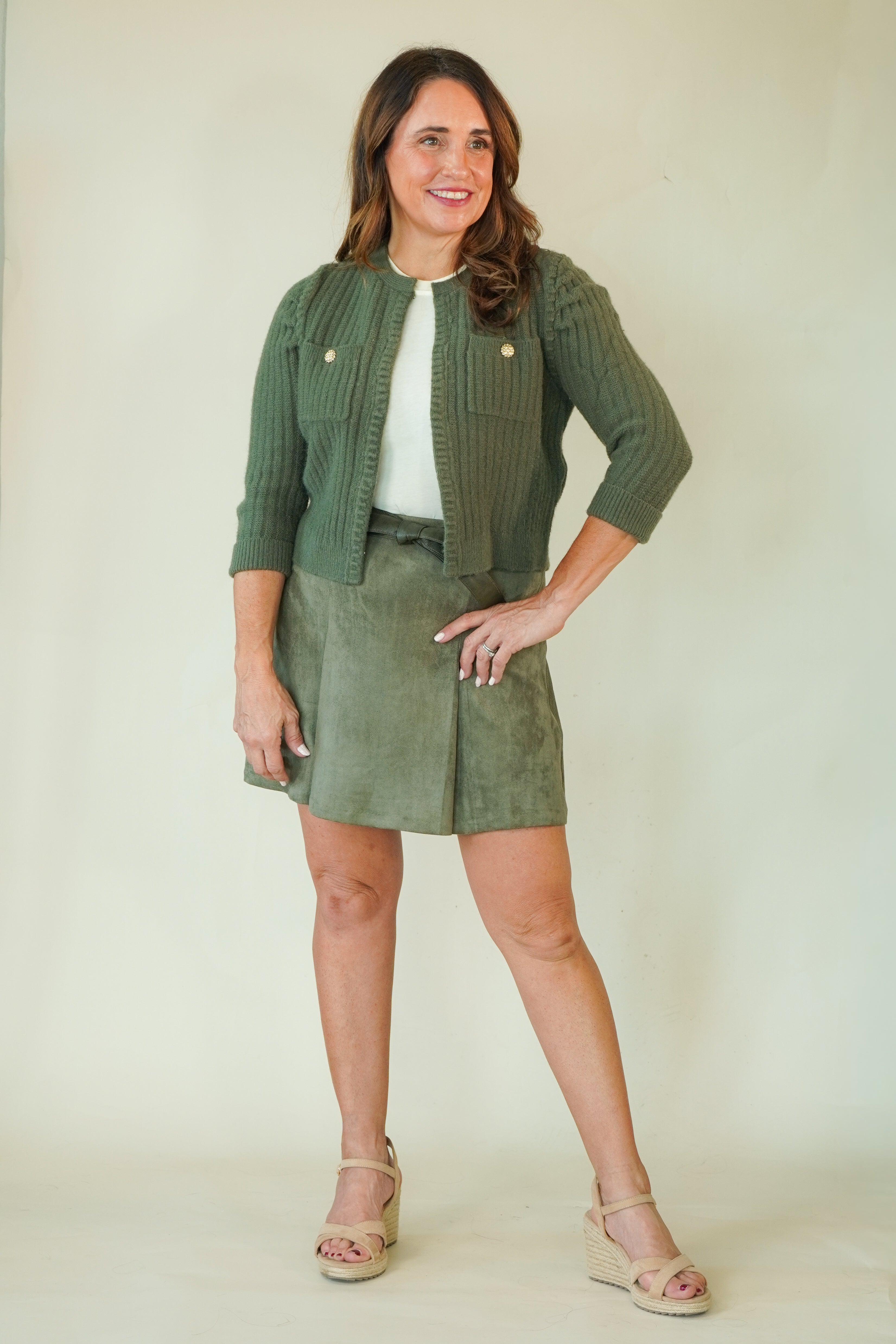 A woman modeling a matching olive green knit cardigan and skirt set with a white top and beige wedge sandals, standing against a plain cream studio backdrop.
