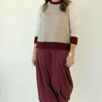 A woman modeling a knit sweater vest with cream and burgundy color blocking, paired with loose burgundy corduroy pants and white sneakers, set against a plain white studio backdrop.