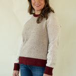 A woman modeling a knit raglan sweater with a speckled beige body, white sleeves, and dark red ribbed trim at the crew neck, cuffs, and hem, paired with blue jeans against a plain cream background.