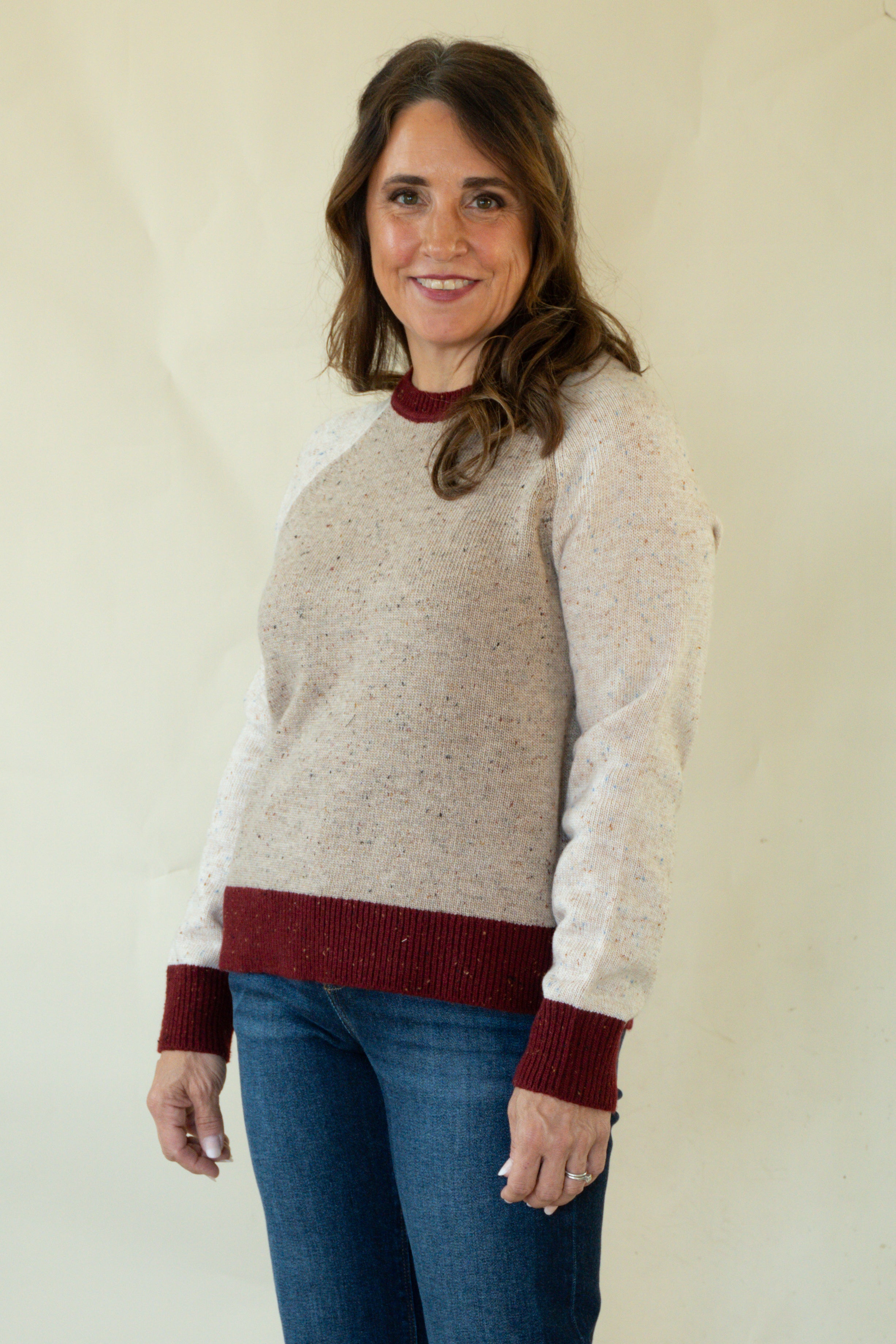 A woman modeling a knit raglan sweater with a speckled beige body, white sleeves, and dark red ribbed trim at the crew neck, cuffs, and hem, paired with blue jeans against a plain cream background.