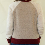 A woman viewed from behind wearing a cream and burgundy knit sweater with heathered back panels and ribbed collar, cuffs, and hem, paired with blue jeans against a plain light background.