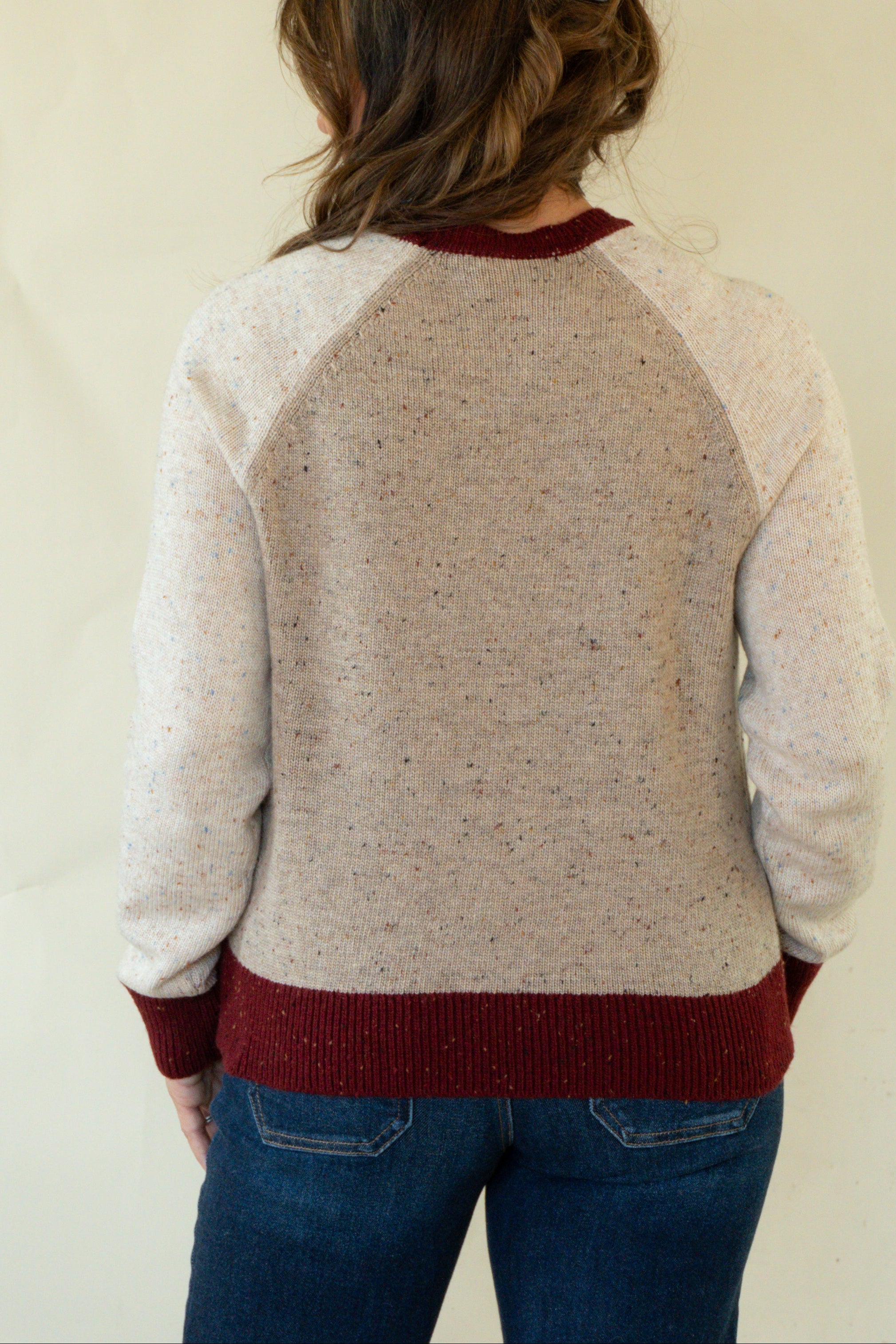 A woman viewed from behind wearing a cream and burgundy knit sweater with heathered back panels and ribbed collar, cuffs, and hem, paired with blue jeans against a plain light background.