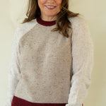 A woman wearing a speckled cream knit sweater with burgundy ribbed collar, cuffs, and hem, paired with blue jeans, standing against a plain beige background.
