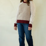 A woman modeling a raglan-style knit sweater with a light beige body and dark burgundy sleeves, collar, and hem, paired with blue jeans and tan boots against a plain cream backdrop.