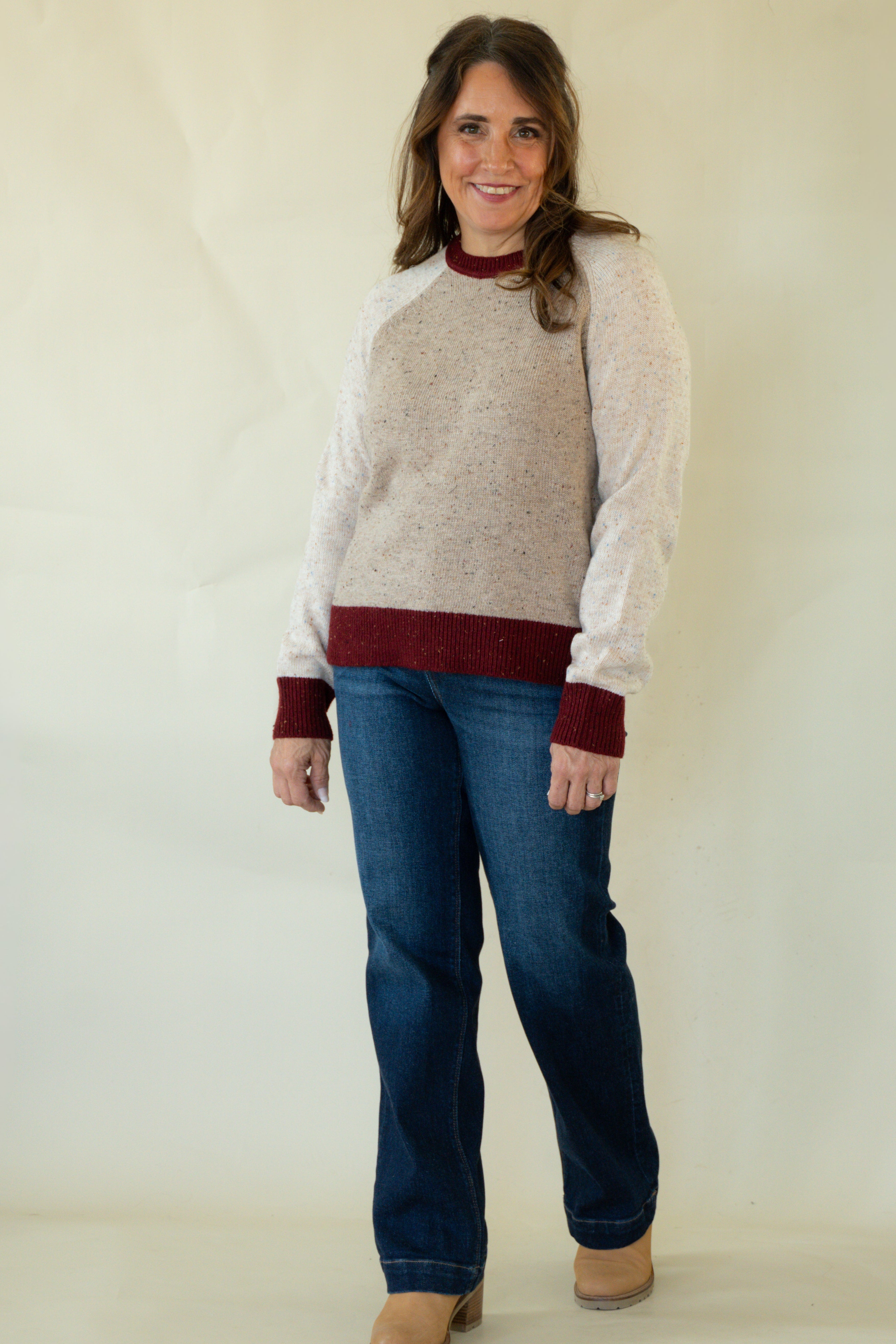 A woman modeling a raglan-style knit sweater with a light beige body and dark burgundy sleeves, collar, and hem, paired with blue jeans and tan boots against a plain cream backdrop.