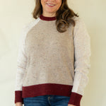 A woman modeling a knit sweater with a speckled beige body, solid white sleeves, and dark burgundy ribbed trim at the crew neck, cuffs, and hem, paired with blue jeans against a plain cream background.