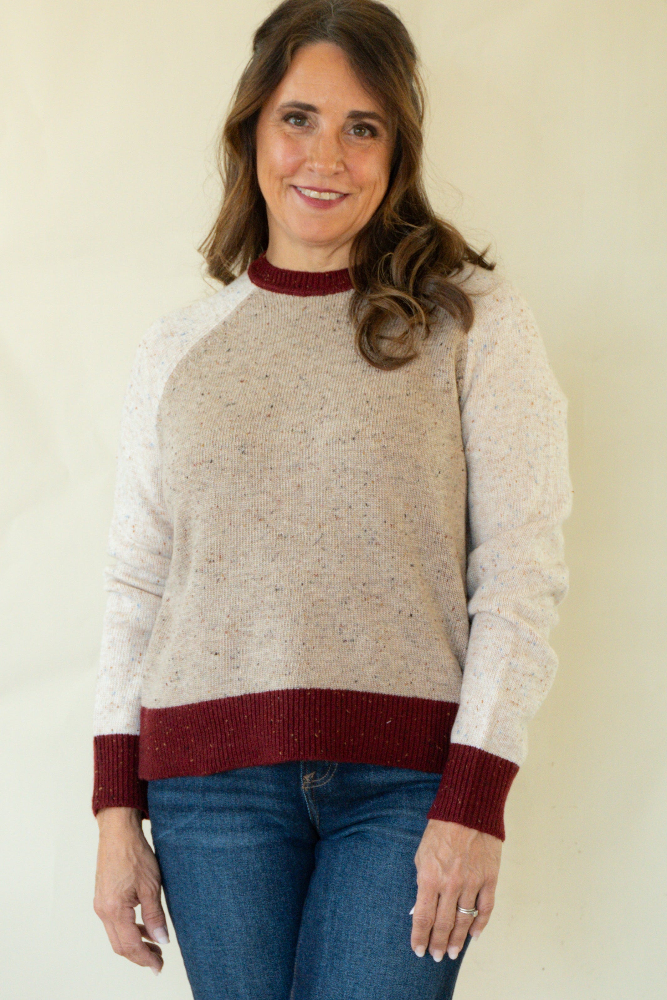 A woman modeling a knit sweater with a speckled beige body, solid white sleeves, and dark burgundy ribbed trim at the crew neck, cuffs, and hem, paired with blue jeans against a plain cream background.