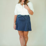 A woman modeling a white short-sleeve collared blouse and a navy blue wrap-style mini skirt with a tie waist, paired with beige strappy heels, against a plain light beige studio background.