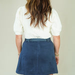 A woman viewed from behind wearing a white short-sleeve puff-sleeve top and a blue suede A-line mini skirt, standing against a plain light beige background.