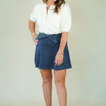 A woman modeling a white short-sleeve blouse and a navy blue wrap-style mini skirt, paired with beige wedge sandals, against a plain light beige studio background.