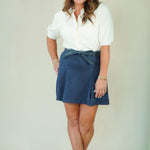 A woman modeling a white short-sleeve button-up shirt and a navy blue skirt with a matching fabric tie belt, paired with beige flat sandals, against a plain light beige studio background.