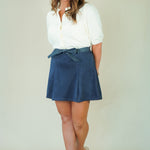 A woman modeling a white short-sleeve button-up shirt and a navy blue pleated mini skirt with a matching tie belt, paired with beige wedge sandals, against a plain light beige studio background.