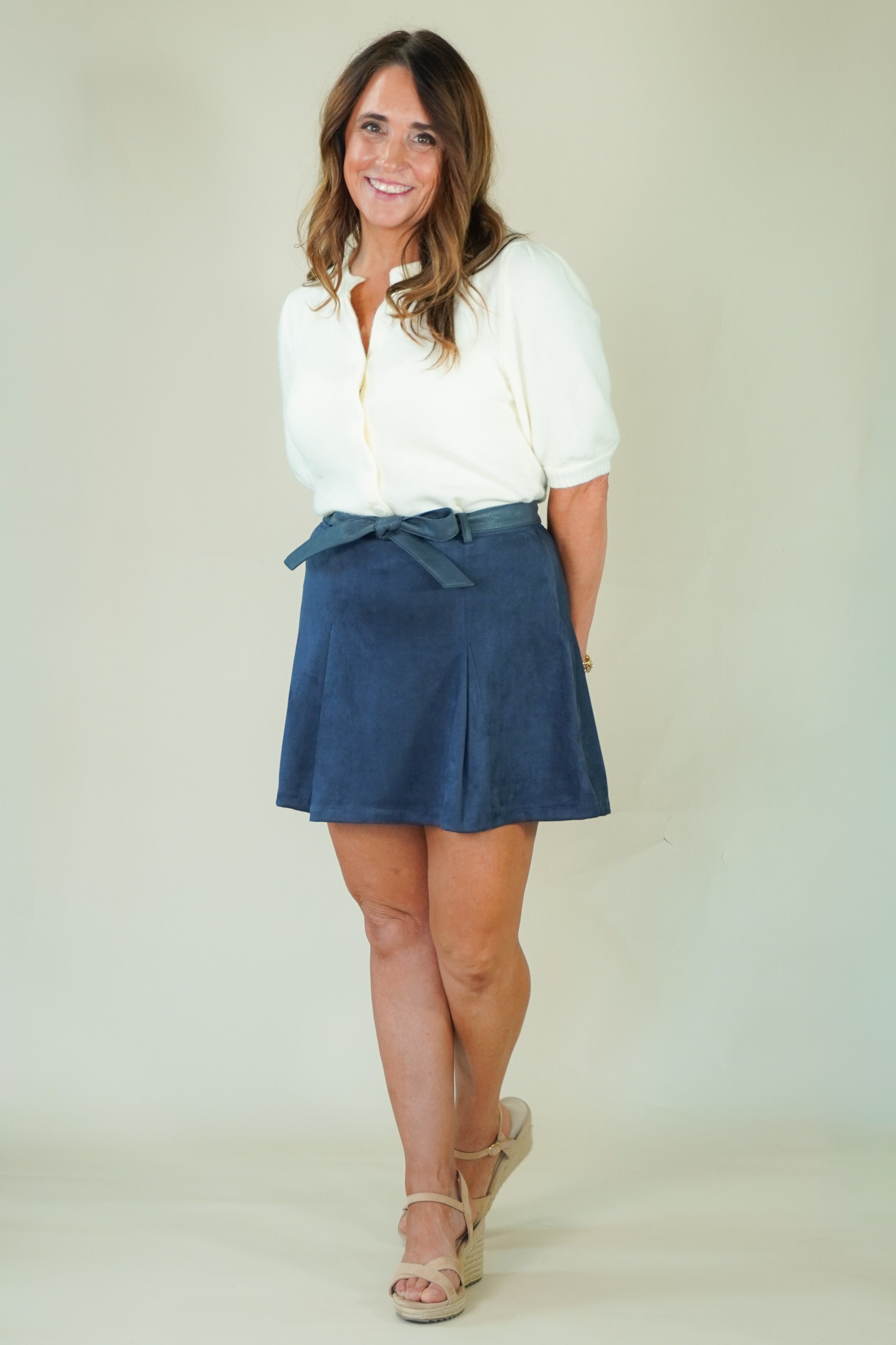A woman modeling a white short-sleeve button-up shirt and a navy blue pleated mini skirt with a matching tie belt, paired with beige wedge sandals, against a plain light beige studio background.