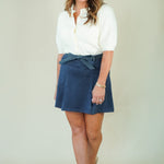 A woman modeling a white short-sleeve blouse and a navy blue wrap-style mini skirt with a tie waist, paired with beige wedge sandals, against a plain light beige studio background.