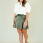 A woman modeling a white long-sleeve blouse and an olive green suede-like mini skirt with a matching belt, paired with beige wedge sandals, against a plain light beige studio background.