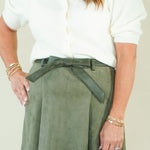 A woman modeling a white short-sleeve blouse and an olive green suede-like mini skirt with a matching belt, paired with beige wedge sandals, against a plain cream studio background.