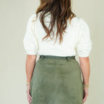 A woman viewed from behind wearing a white short-sleeve top and an olive green mini skirt, standing against a plain light beige studio background.