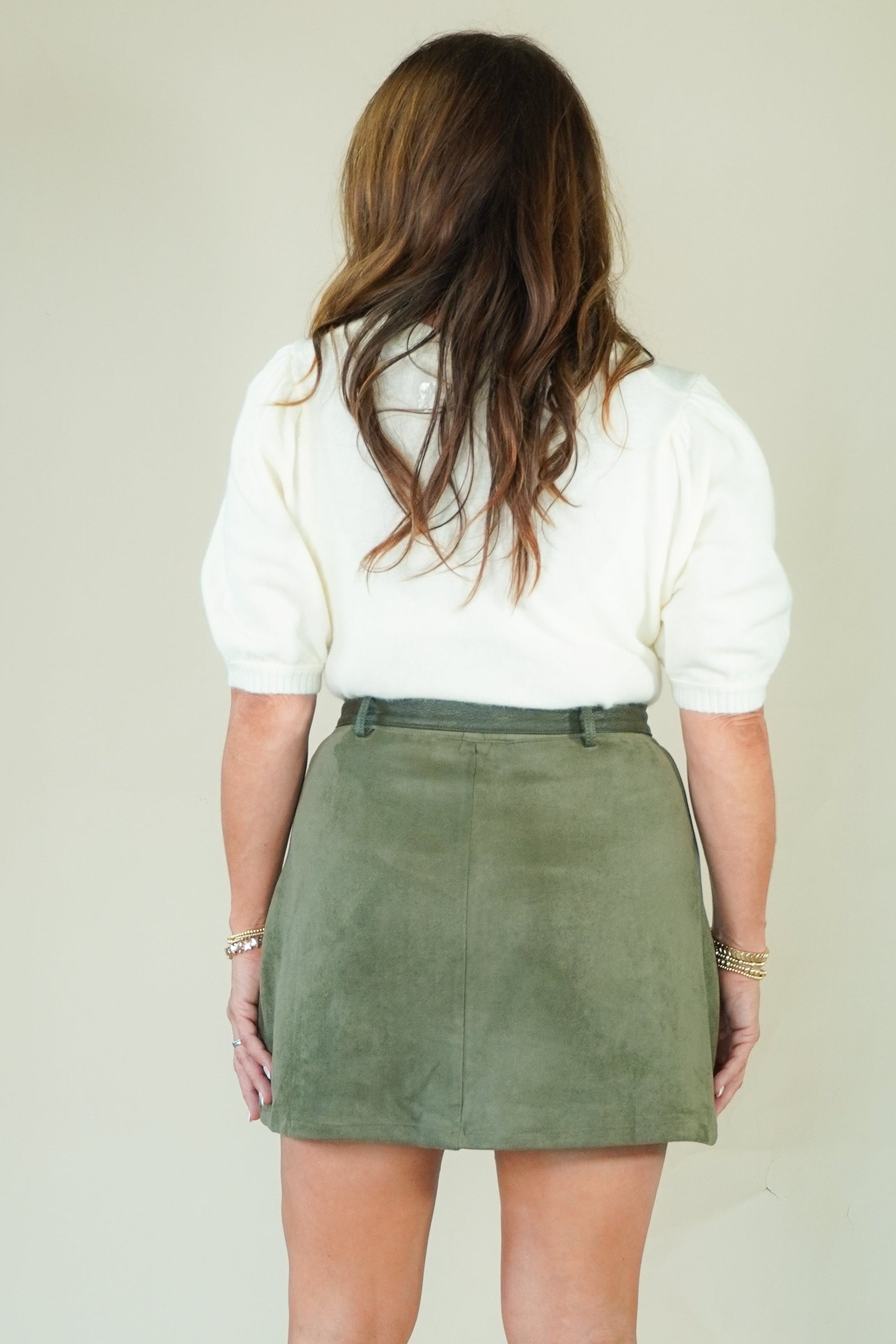 A woman viewed from behind wearing a white short-sleeve top and an olive green mini skirt, standing against a plain light beige studio background.