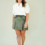 A woman modeling a white long-sleeve blouse and an olive green mini skirt with a matching belt, paired with beige strappy sandals, against a plain cream studio background.