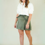 A woman modeling a white long-sleeve blouse and an olive green suede-like mini skirt with a matching belt, paired with beige wedge sandals, against a plain cream studio background.