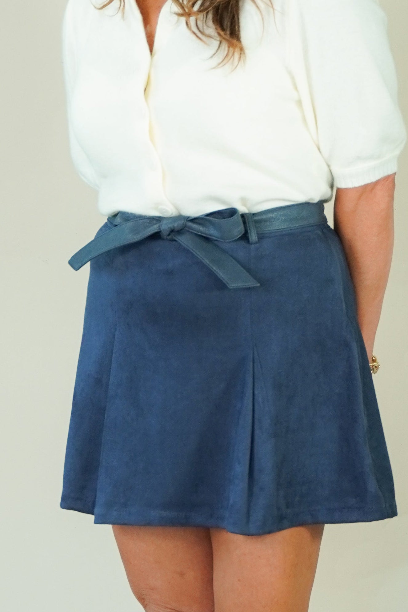 A woman modeling a white short-sleeve button-up shirt and a navy blue pleated mini skirt with a matching tie belt, paired with beige wedge sandals, against a plain light beige studio background.