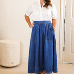 Woman wearing a white shirt and blue skirt standing in a room with a mirror and dresser.