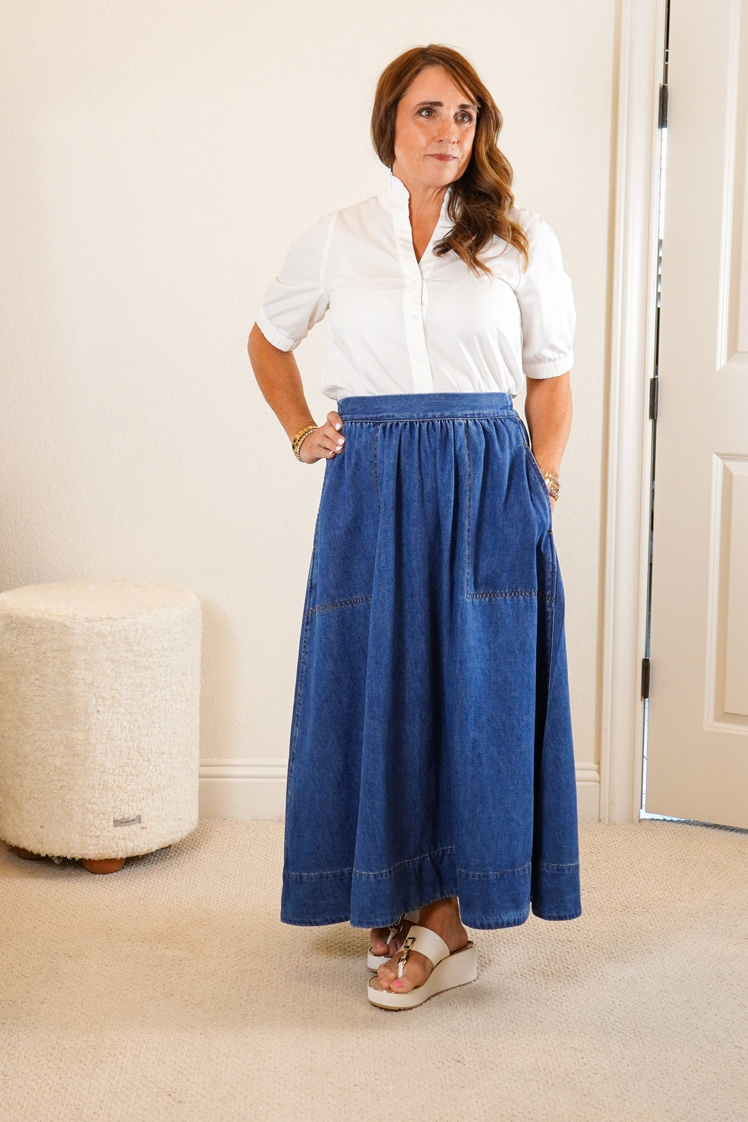 Woman wearing a white shirt and blue skirt standing in a room with a mirror and dresser.