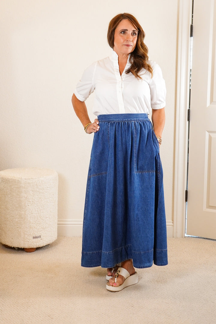 Woman wearing a white shirt and blue skirt standing in a room with a mirror and dresser.