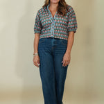 A woman modeling a short-sleeve button-up shirt with a colorful paisley pattern, paired with dark blue jeans and beige wedge sandals, standing against a plain beige studio backdrop.