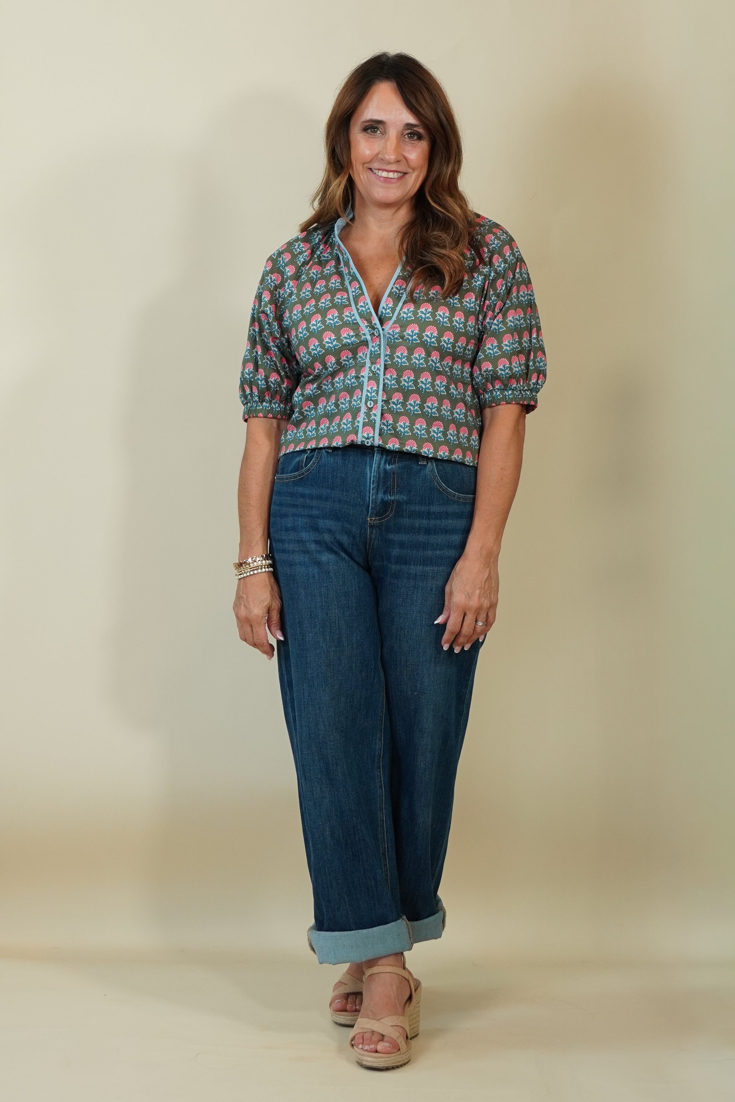 A woman modeling a short-sleeve button-up shirt with a colorful paisley pattern, paired with dark blue jeans and beige wedge sandals, standing against a plain beige studio backdrop.
