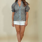 A woman modeling a short-sleeve button-up shirt with a blue, green, and pink elephant print, paired with white shorts and beige sandals, against a solid beige studio background.