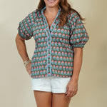 A woman modeling a short-sleeve button-up shirt with a colorful geometric pattern in teal, pink, and white, paired with white shorts and beige sandals, against a solid beige studio background.