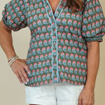 A woman modeling a short-sleeve button-up blouse with an all-over pink, blue, and green paisley pattern, paired with white shorts and beige sandals, against a solid beige studio background.