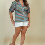 A woman modeling a short-sleeve button-up shirt with a colorful geometric pattern in teal, pink, and white, paired with white shorts and beige wedge sandals, against a solid beige studio background.