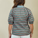 A woman shown from the back wearing a short-sleeve button-up shirt with a repeating green, pink, and white floral pattern, paired with white shorts, against a solid beige background.