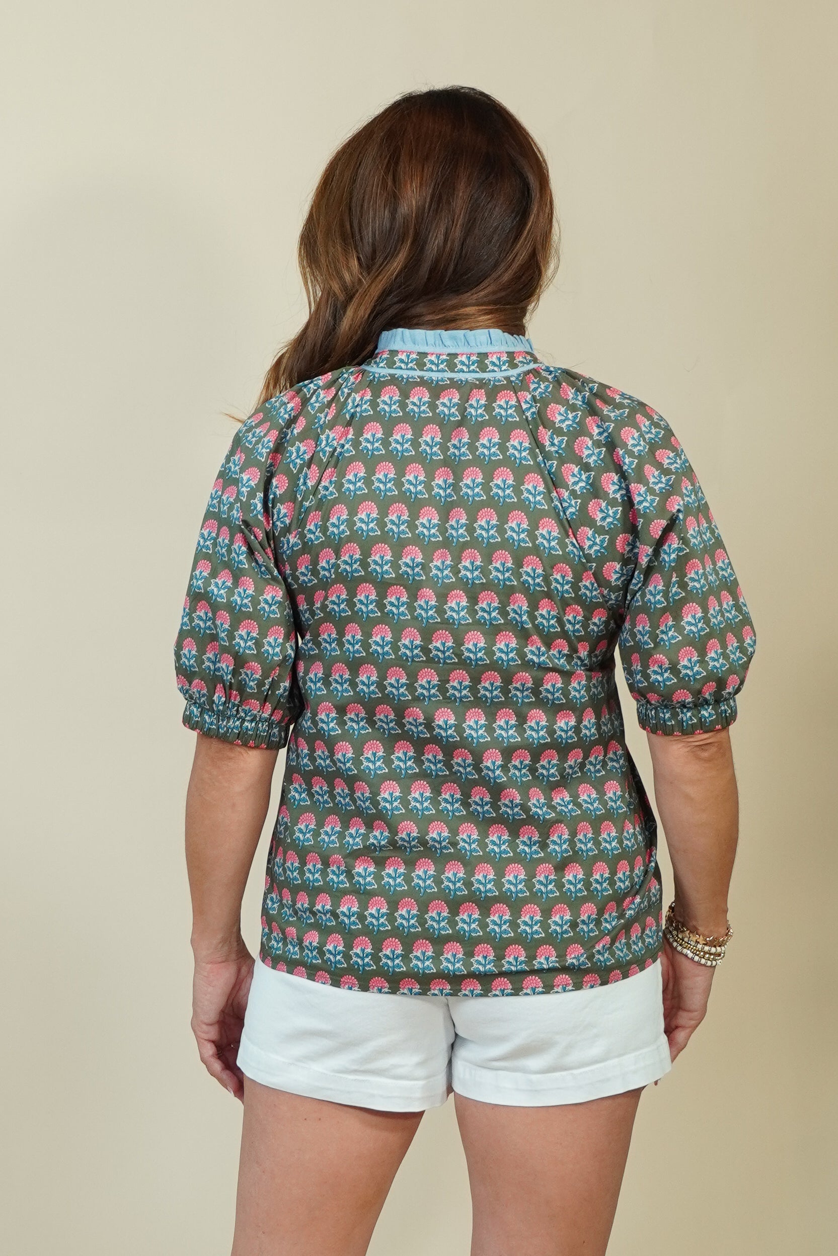 A woman shown from the back wearing a short-sleeve button-up shirt with a repeating green, pink, and white floral pattern, paired with white shorts, against a solid beige background.
