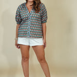 A woman modeling a short-sleeve button-up shirt with a colorful repeating elephant print, paired with white shorts and beige wedge sandals, against a solid beige studio background.