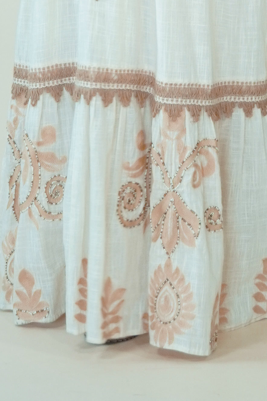 Close-up of a white or off-white fabric garment with a ruffled hem and dusty pink floral embroidery and embroidered border details.