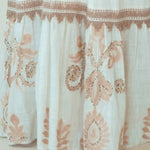 Close-up of a white or off-white fabric garment with a ruffled hem and dusty pink floral embroidery and embroidered border details.