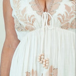 A woman modeling a sleeveless, floor-length white maxi dress with beige floral embroidery and a tie-neck detail, set against a plain cream studio background.
