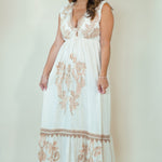A woman modeling a sleeveless white maxi dress with beige floral embroidery, featuring a deep V-neckline and a tiered skirt, set against a plain cream studio background.