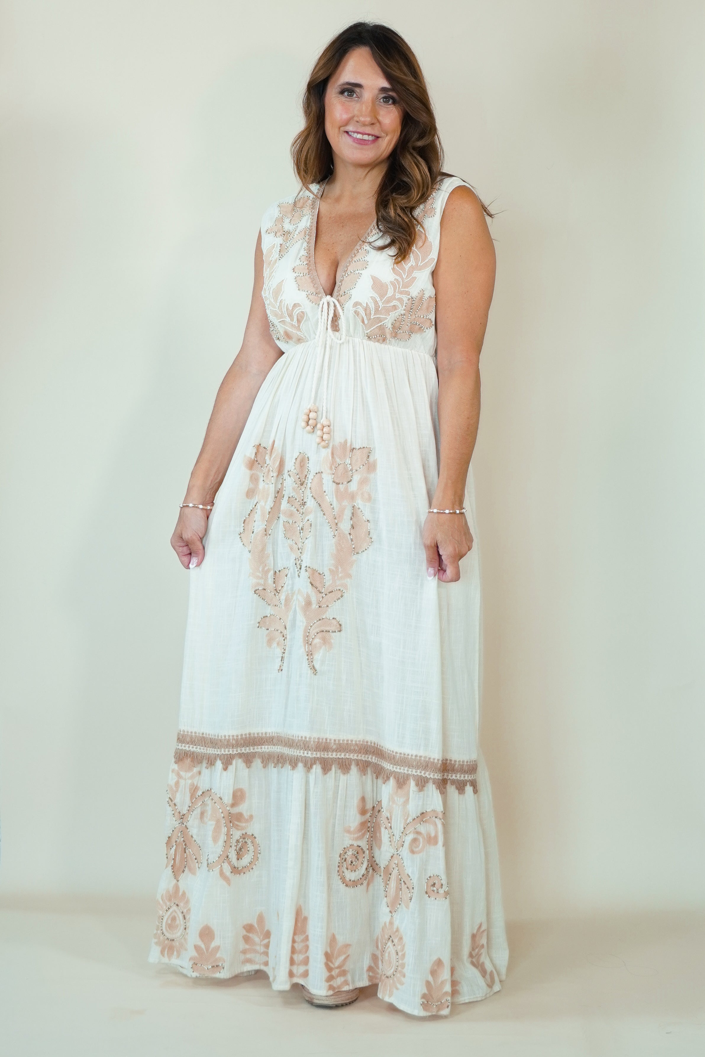 A woman modeling a sleeveless white maxi dress with beige floral embroidery, featuring a deep V-neckline and a tiered skirt, set against a plain cream studio background.