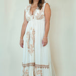 A woman modeling a sleeveless, floor-length white maxi dress with beige floral embroidery and a tie-neck detail, set against a plain cream studio backdrop.