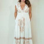 A woman modeling a sleeveless white maxi dress with beige floral embroidery, a deep V-neckline, and side pockets, set against a plain off-white studio background.
