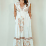 A woman modeling a sleeveless white maxi dress with beige floral embroidery and a deep V-neckline, standing against a plain beige studio background.