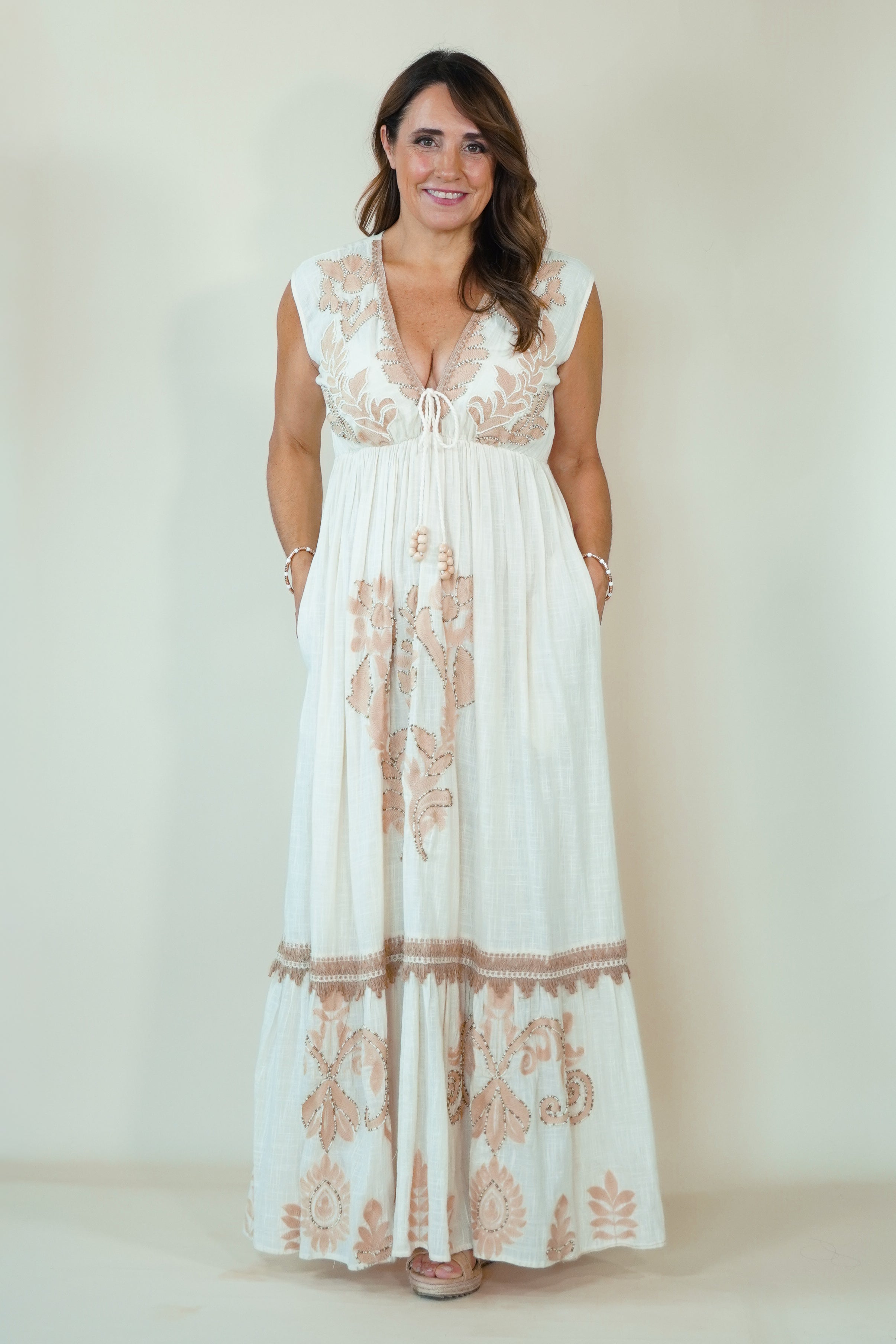 A woman modeling a sleeveless white maxi dress with beige floral embroidery and a deep V-neckline, standing against a plain beige studio background.