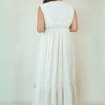 A woman viewed from behind wearing a sleeveless, tiered white maxi dress against a plain, warm off-white studio background.